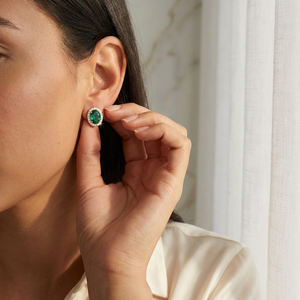 Model wearing Lab-Grown Emerald Halo Stud Earrings with Diamonds, showcasing elegance and vibrant color next to a sheer curtain.