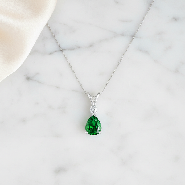 Prong-Set Lab-Grown Emerald Teardrop V-Bale Pendant with Diamond on marble background.
