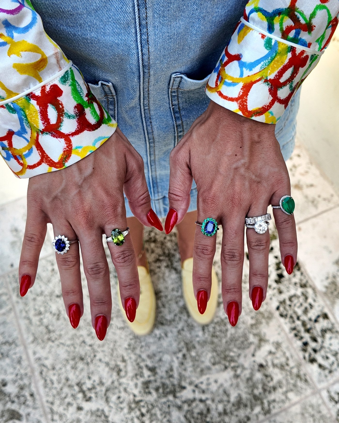 Six Irdoja rings worn across two hands - blue sapphire, peridot, an emerald-cut blue stone with a green halo, a diamond eternity band, a round solitaire and a green pear halo - against a painted silk shirt and denim.